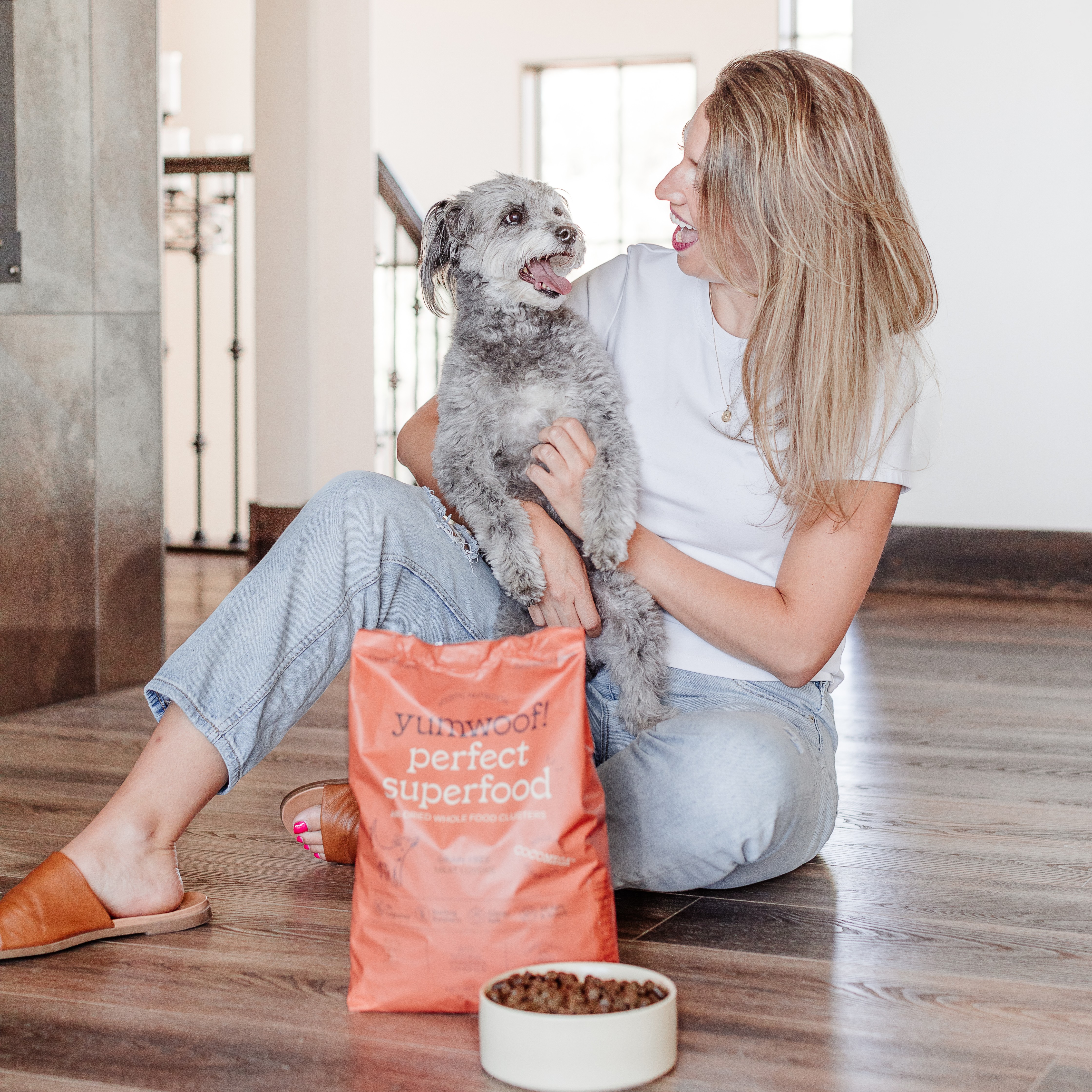A smiling woman sits on the floor, holding her small grey dog next to a bag and bowl of dog food.