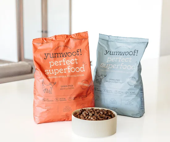 Two bags of dog food labeled 'yumwoof! perfect superfood' beside a bowl of kibble.