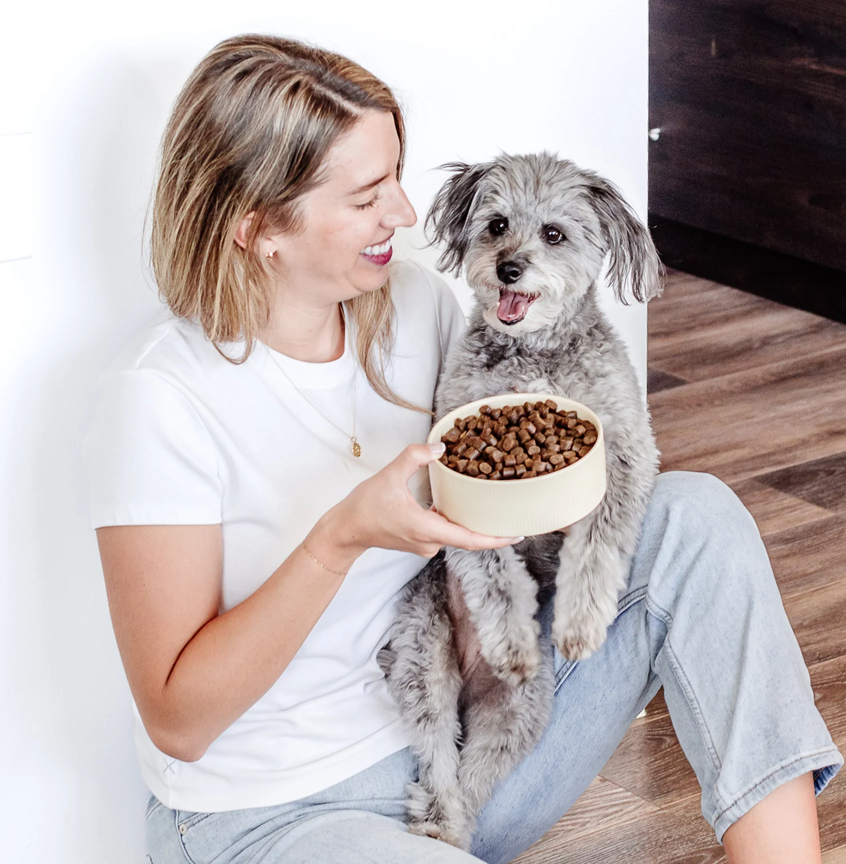A woman sits on the floor, holding a small gray dog and a bowl of dog food.