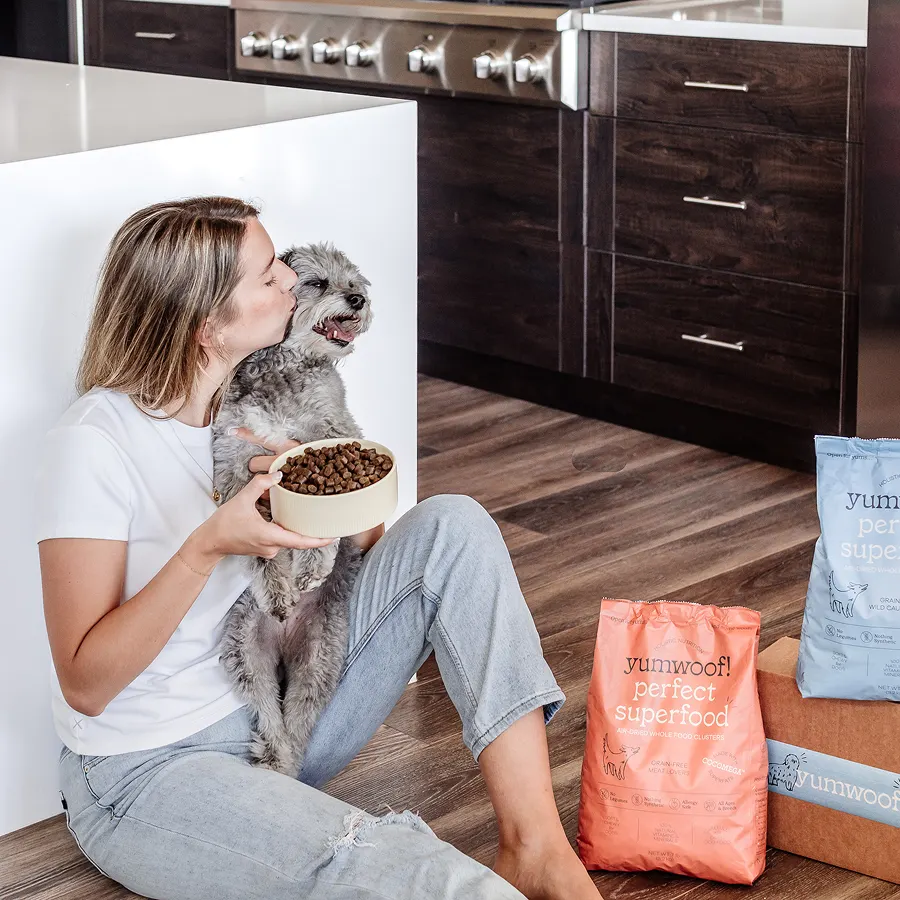 Person hugging a gray dog while holding a bowl of dog food in a kitchen.