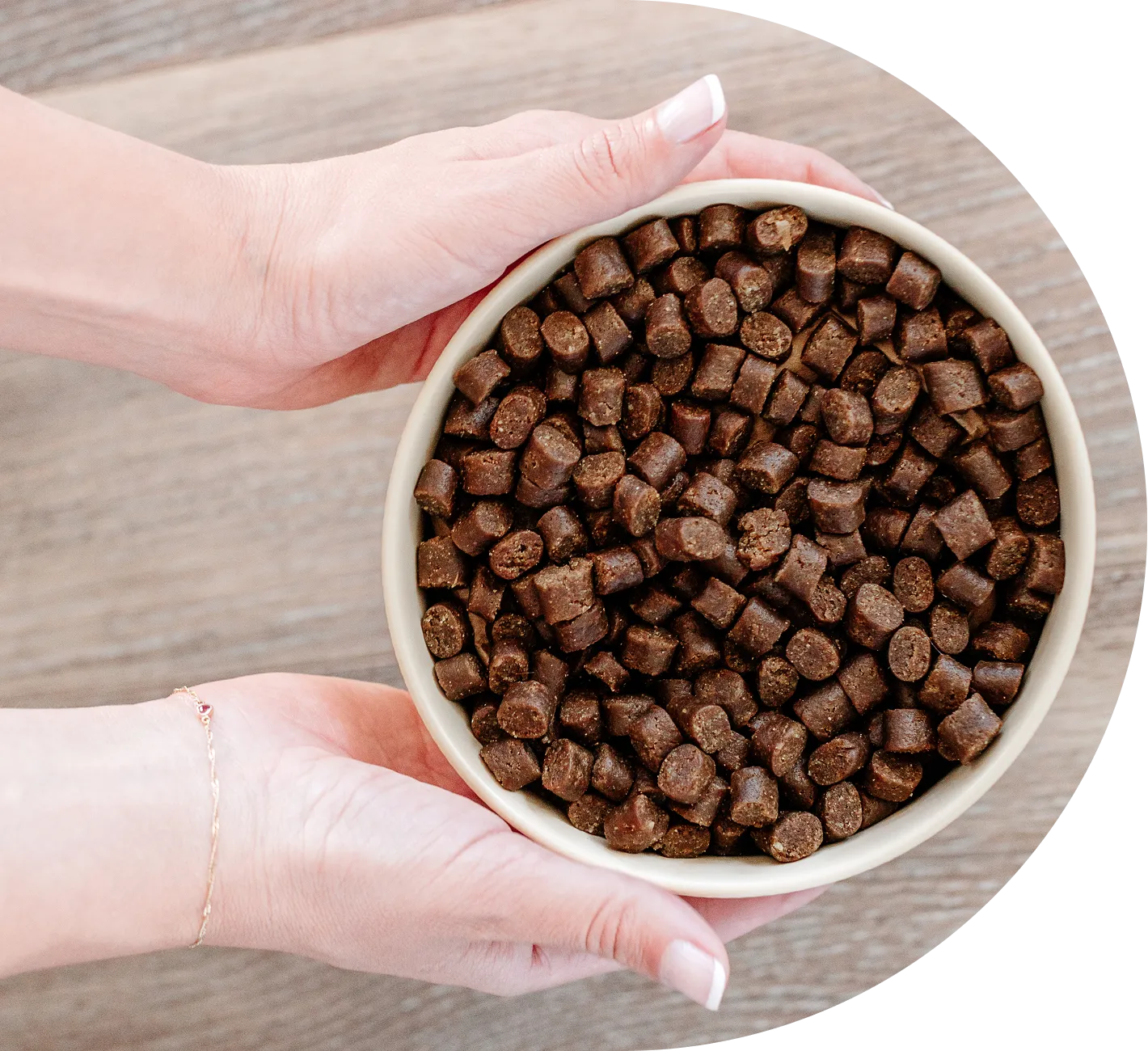 A pair of hands holding a white bowl filled with brown, cylindrical pet food kibble over a wooden surface.