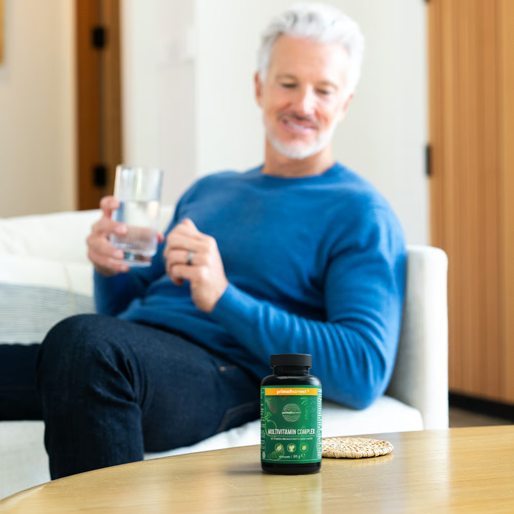 A bottle of multivitamins on a table, with a man holding a glass of water in the background.