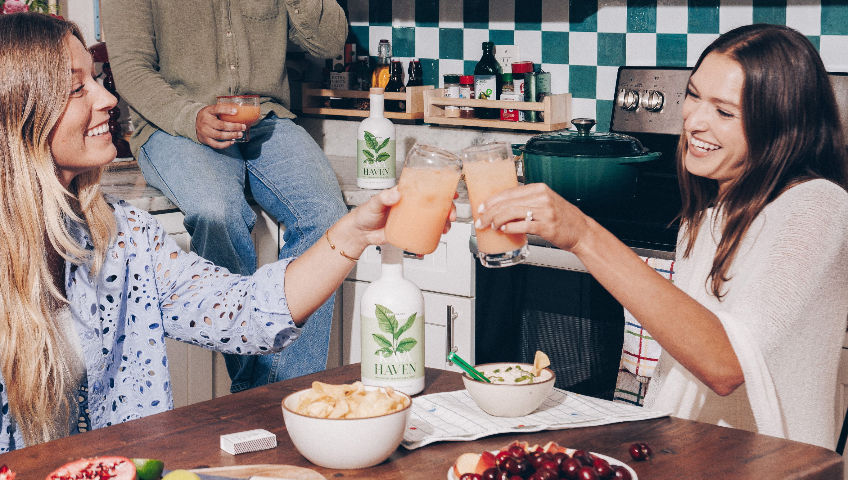 Three people smiling and toasting with drinks in a kitchen filled with snacks and a bottle of Haven.