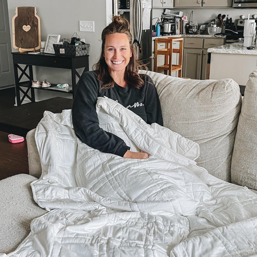 Person smiling while sitting wrapped in a white blanket on a couch in a cozy living room.