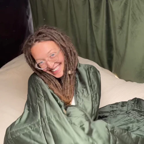 Smiling person with glasses wrapped in a green blanket on a bed.