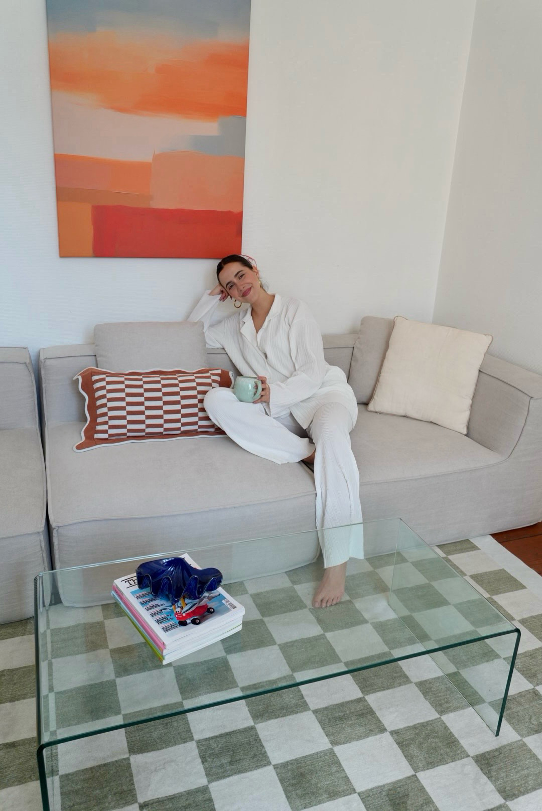 Person in white outfit sitting on a sofa with a mug, abstract painting behind.