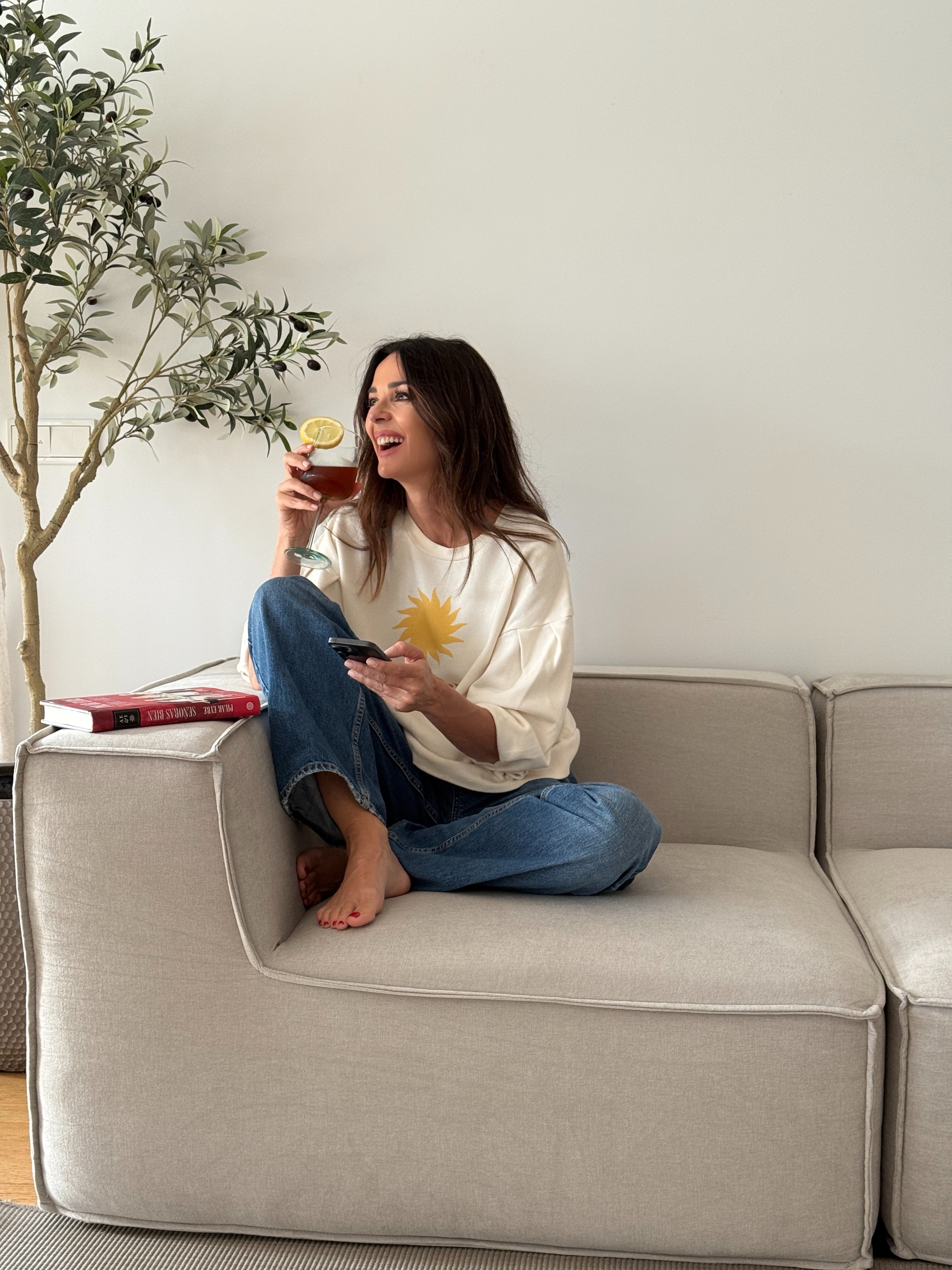 Woman sitting on a couch, holding a drink with a lemon slice, next to a potted plant.