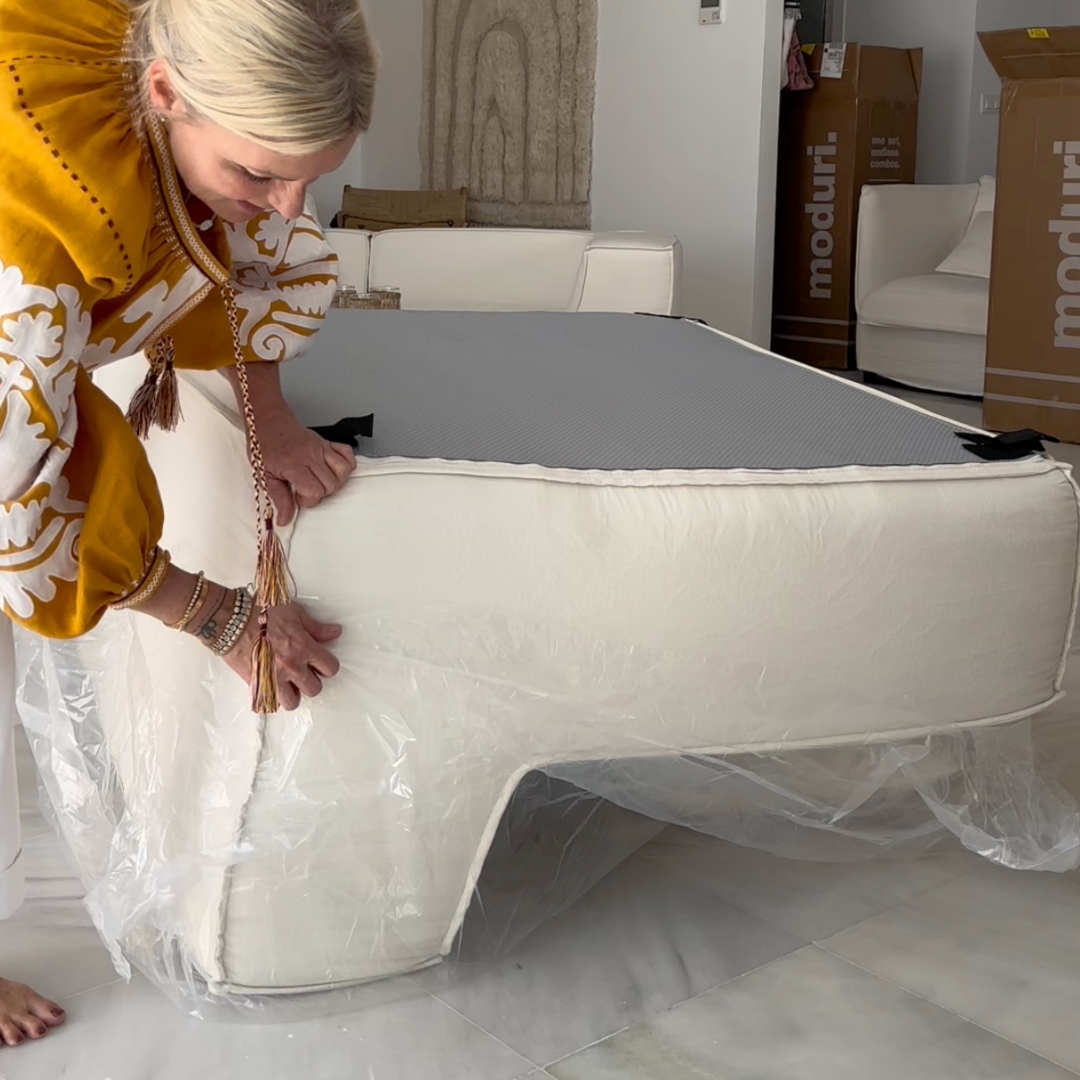 Person assembling a white couch with plastic wrapping in a living room.