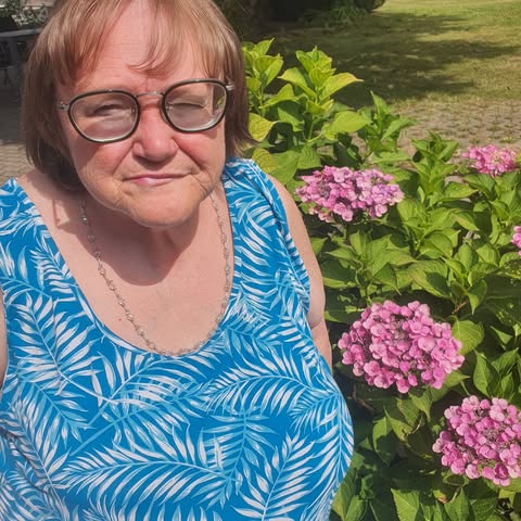 An older woman with glasses wearing a blue leaf-print top stands next to a pink hydrangea bush.