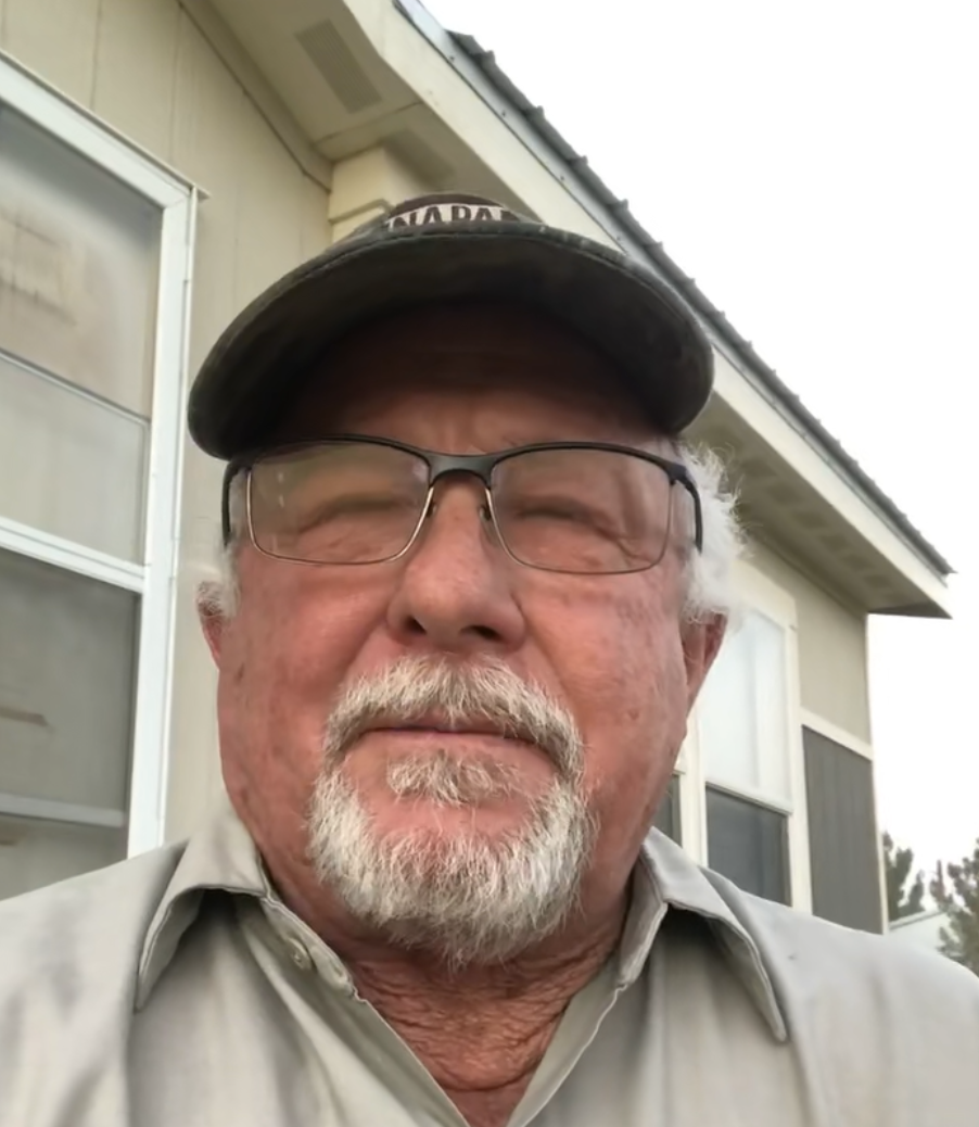 Man with glasses and cap standing outside a house.