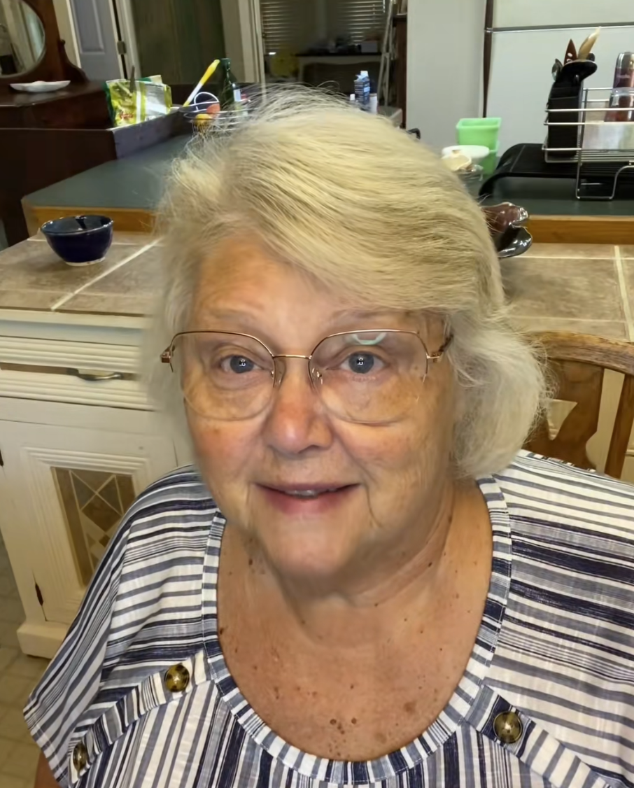 Elderly woman with glasses smiling in a kitchen setting.