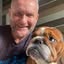 A close-up photo of a man with a shaved head smiling next to a bulldog.