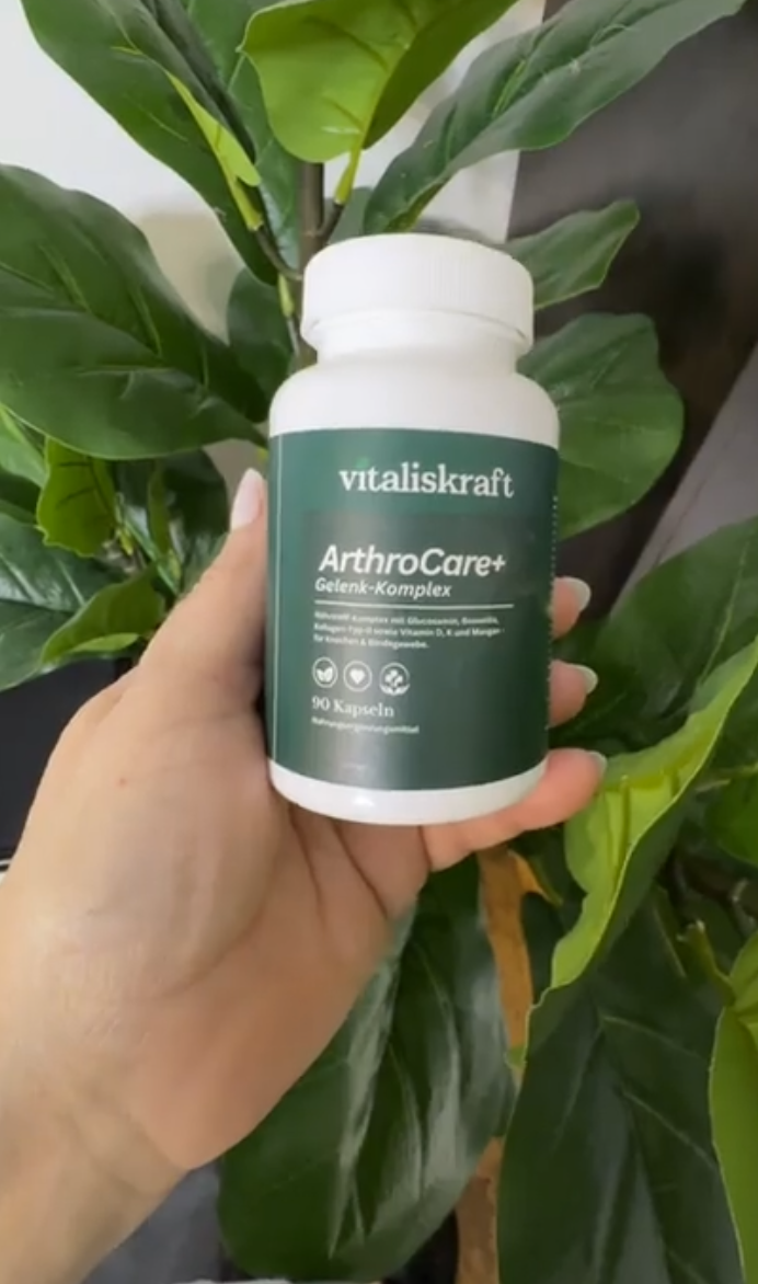 A hand holding a bottle of Vitaliskraft ArthroCare+ supplements in front of a large green-leafed plant.