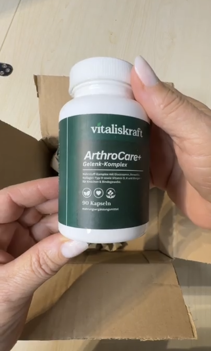 A person's hands holding a white bottle of Vitaliskraft ArthroCare+ supplements over a cardboard box.