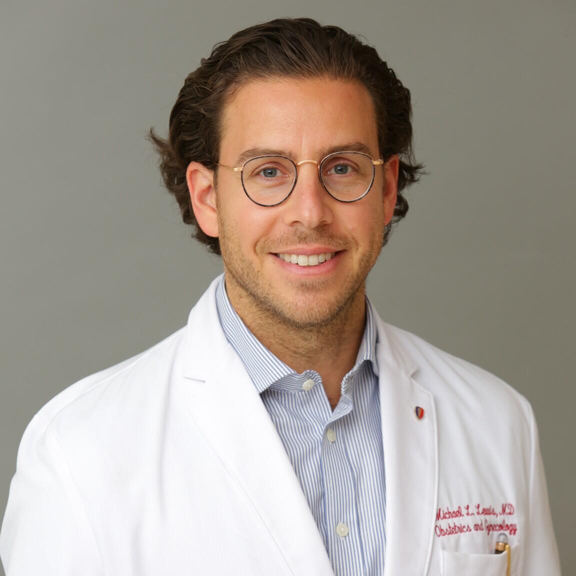 Person in a white coat with glasses, smiling against a gray background.