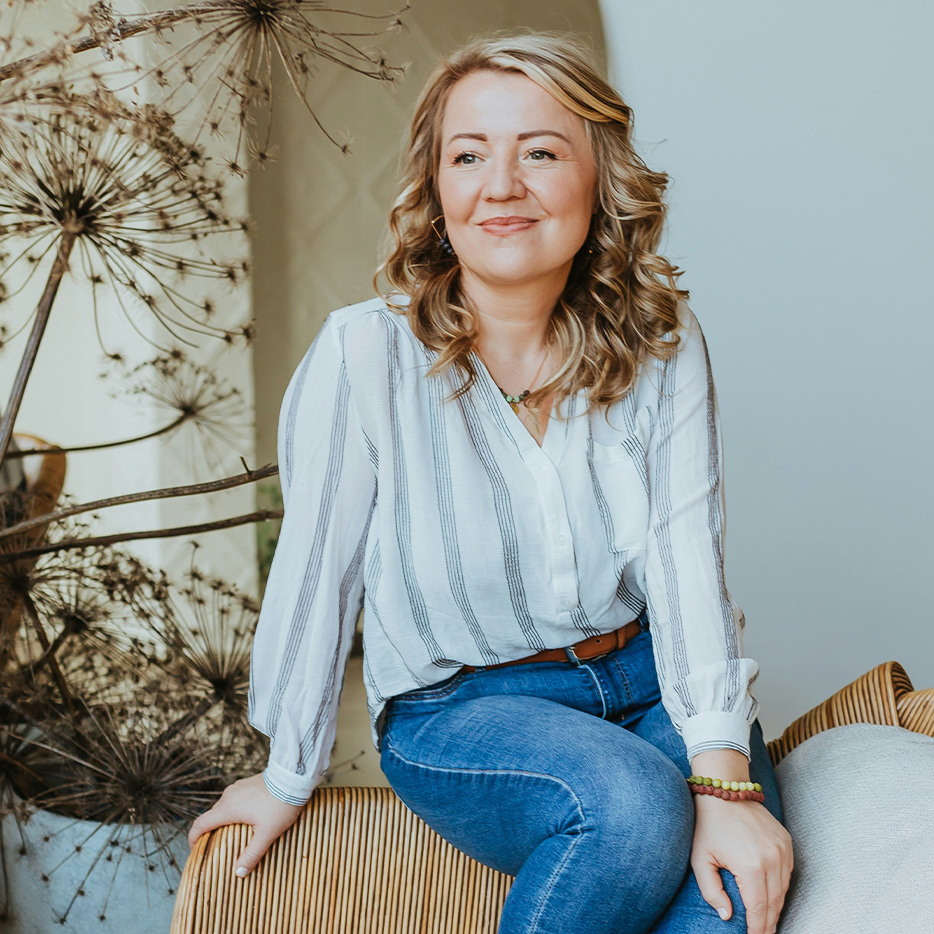 Woman sitting on a chair, smiling, with dried plants in the background.