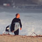 Person in wetsuit holding a surfboard, standing on a beach with waves in the background.