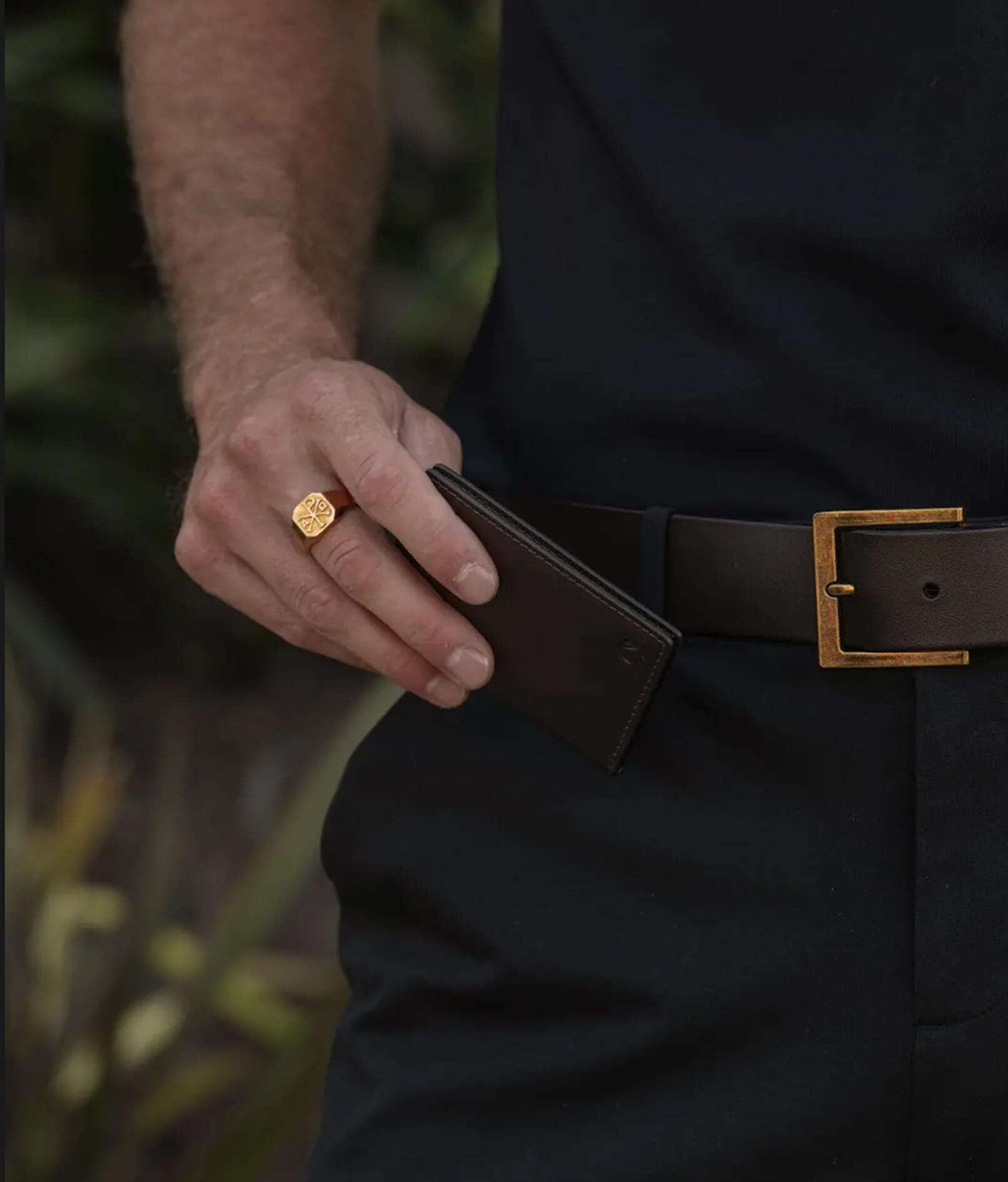 A person holding a black wallet, wearing a ring and brown belt.