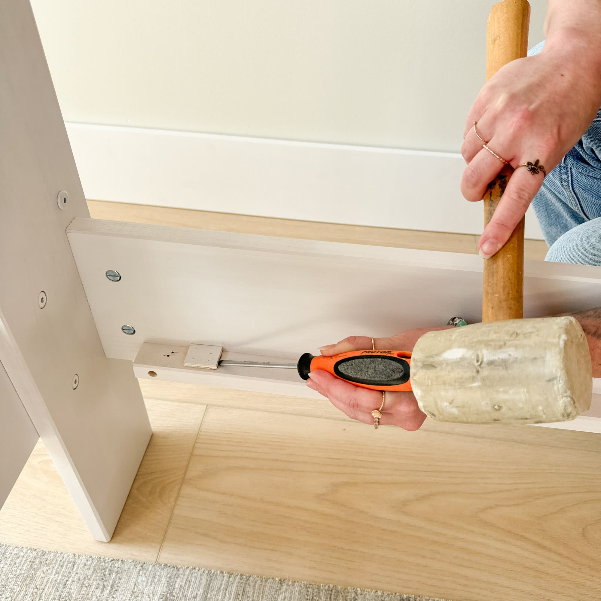 A person using a wooden mallet and a screwdriver to pry off a small block of wood from a bed frame.