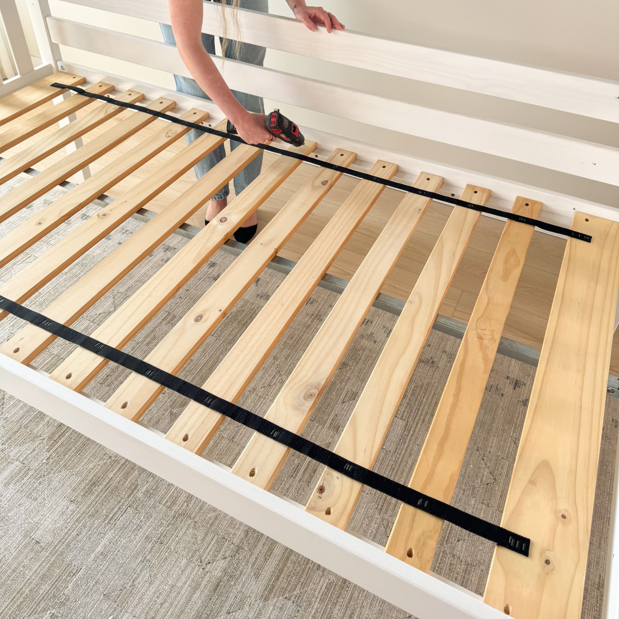 A person using a power tool to screw in wooden slats into a bed frame.