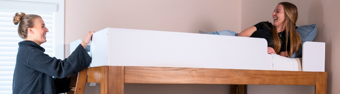 Three friends relaxing on a wooden loft bed with ladder in a cozy college-dorm-style bedroom — top bunk mattress and lower full bed with pillows.