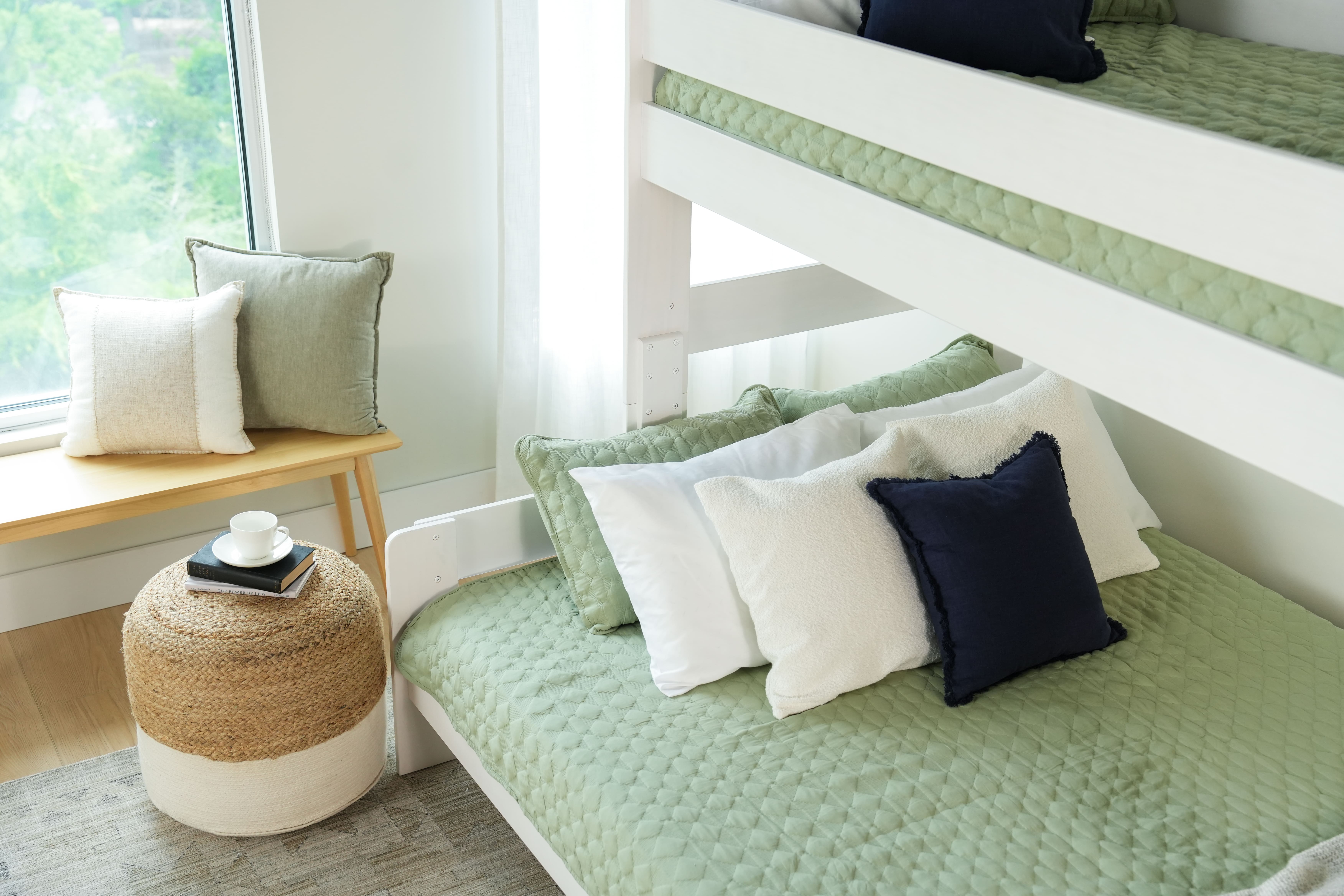 A white bunk bed with green bedding with an ottoman and bench nearby.