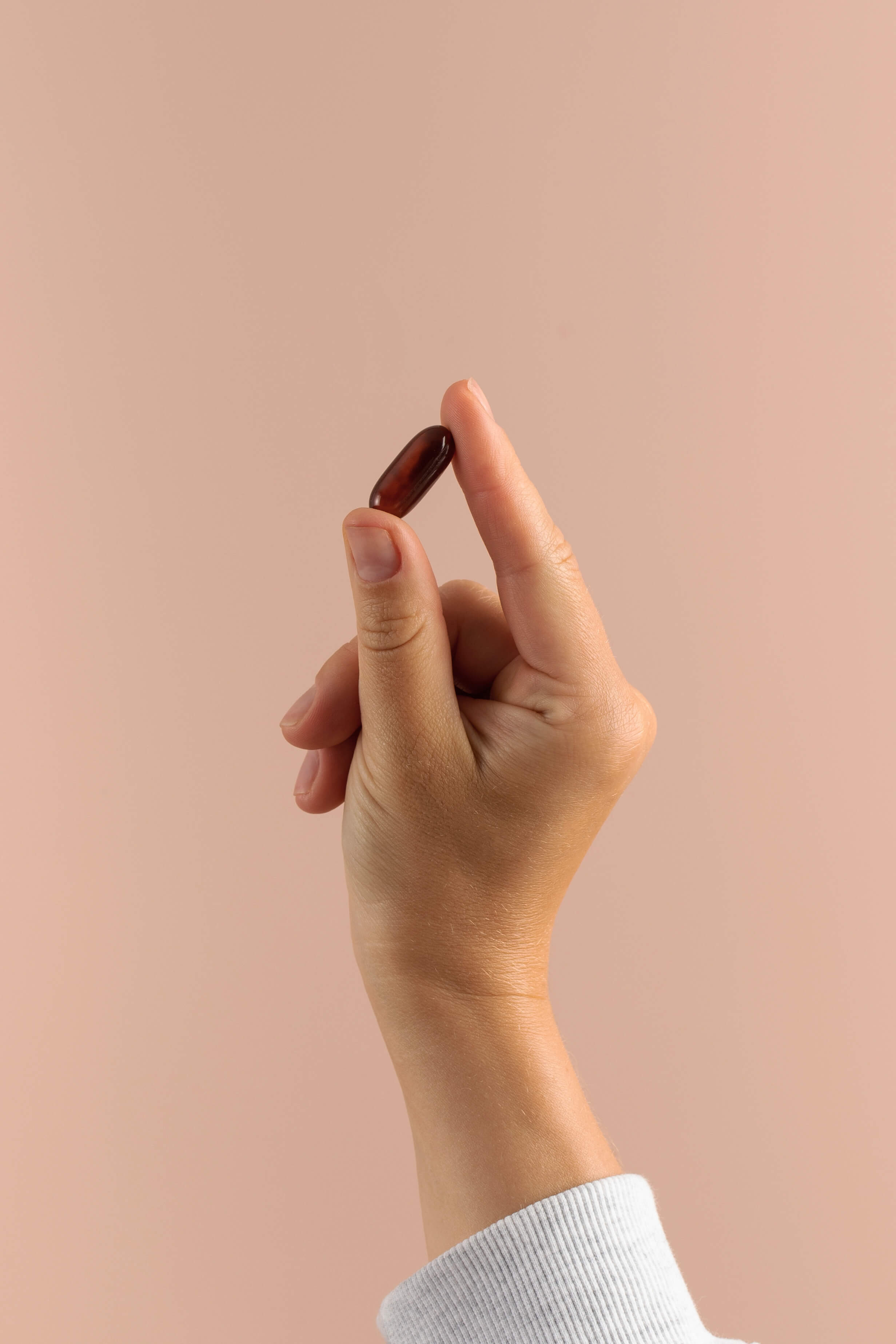 Hand holding a small, brown gel capsule against a beige background.