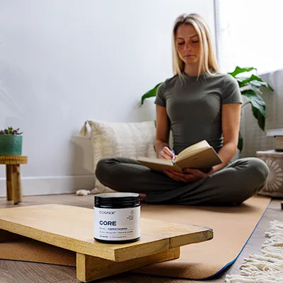 Woman sitting cross-legged, writing in a notebook, with a supplement container in the foreground.