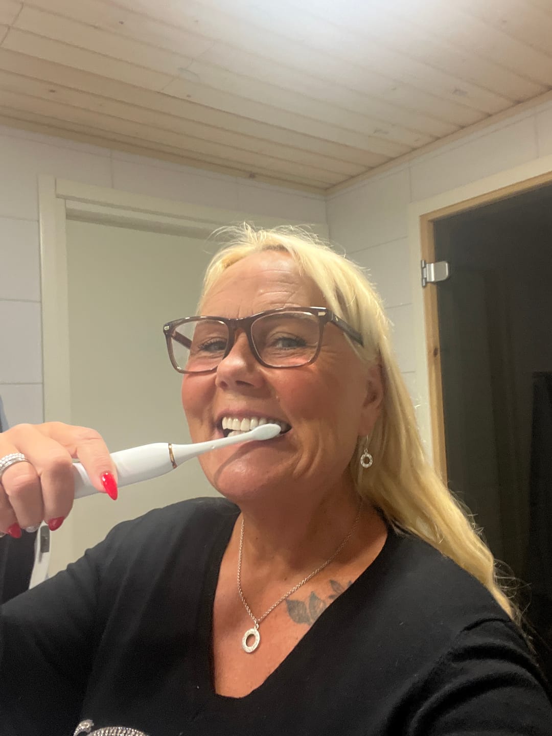 A smiling blonde woman with glasses brushes her teeth with a white electric toothbrush in a bathroom.