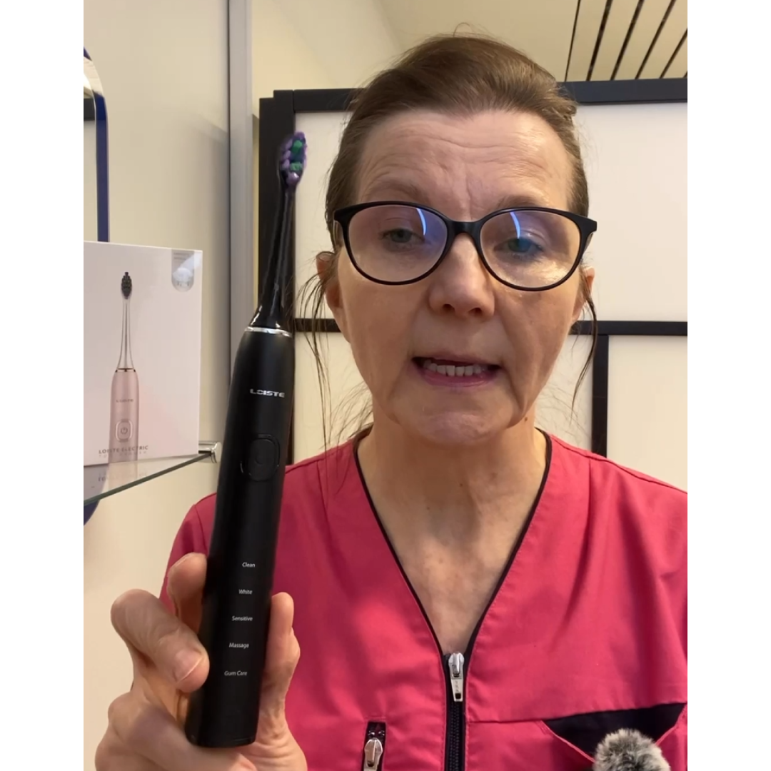 Person holding a black electric toothbrush, wearing glasses and a pink top.