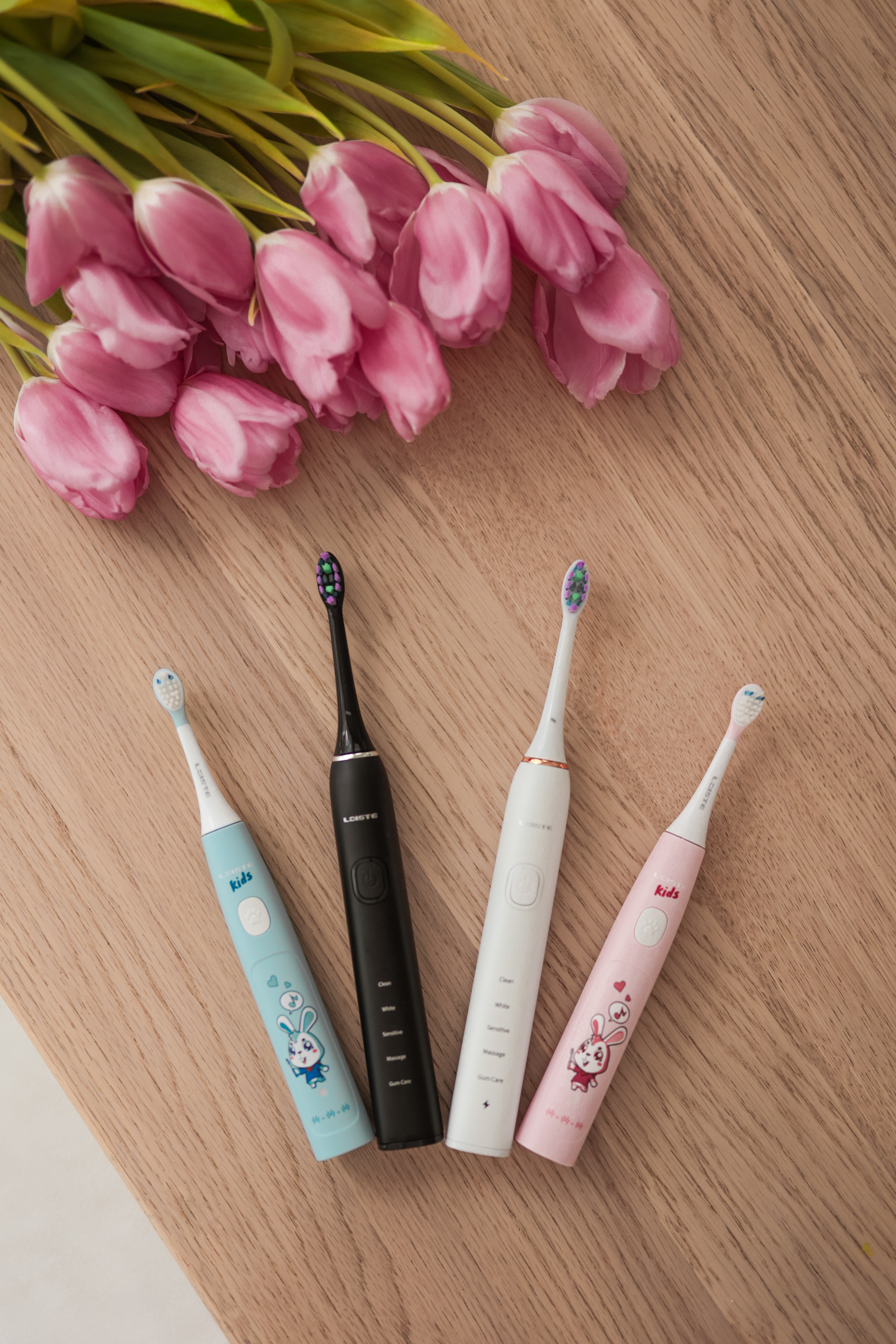 Four electric toothbrushes on a table beside pink tulips.