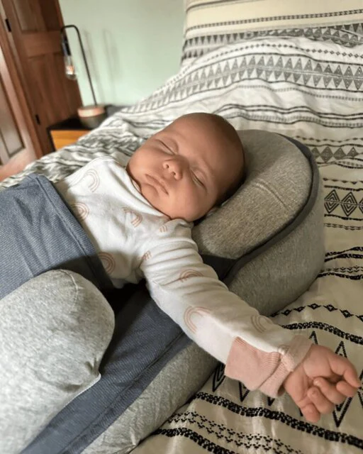 A newborn baby wearing a rainbow-patterned onesie sleeps soundly on a grey support pillow on a bed.