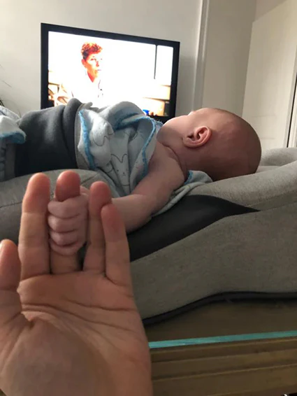 A newborn baby lies on its back, holding an adult's finger in front of a television.