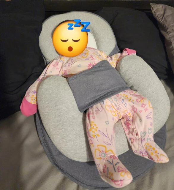 A baby in pink floral pajamas rests in a gray lounger, with a sleeping emoji covering its face.