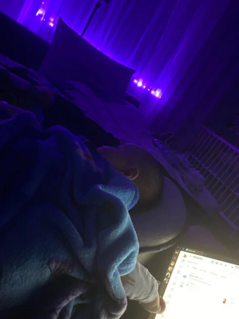 A baby sleeps under a blue blanket in a dark room lit by purple lights and a laptop screen.
