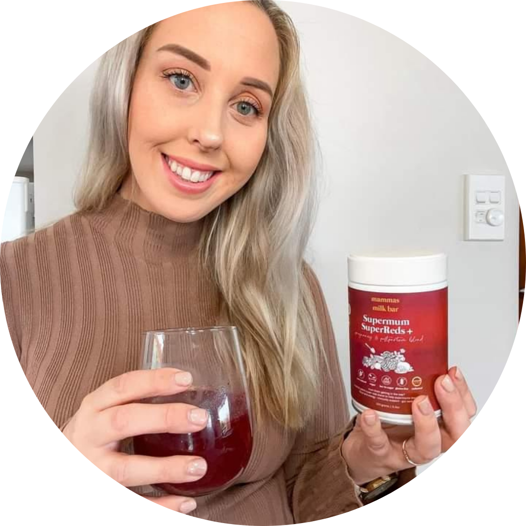 A smiling blonde woman holds a glass of red liquid and a container of Supermum SuperReds+.