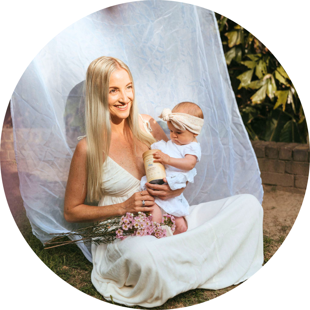 A smiling blonde woman in a white dress sits on the grass, holding a baby on her lap.