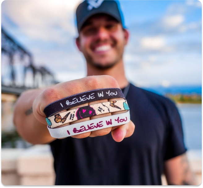 Person holding wristbands with 'I BELIEVE IN YOU' text, smiling in the background.