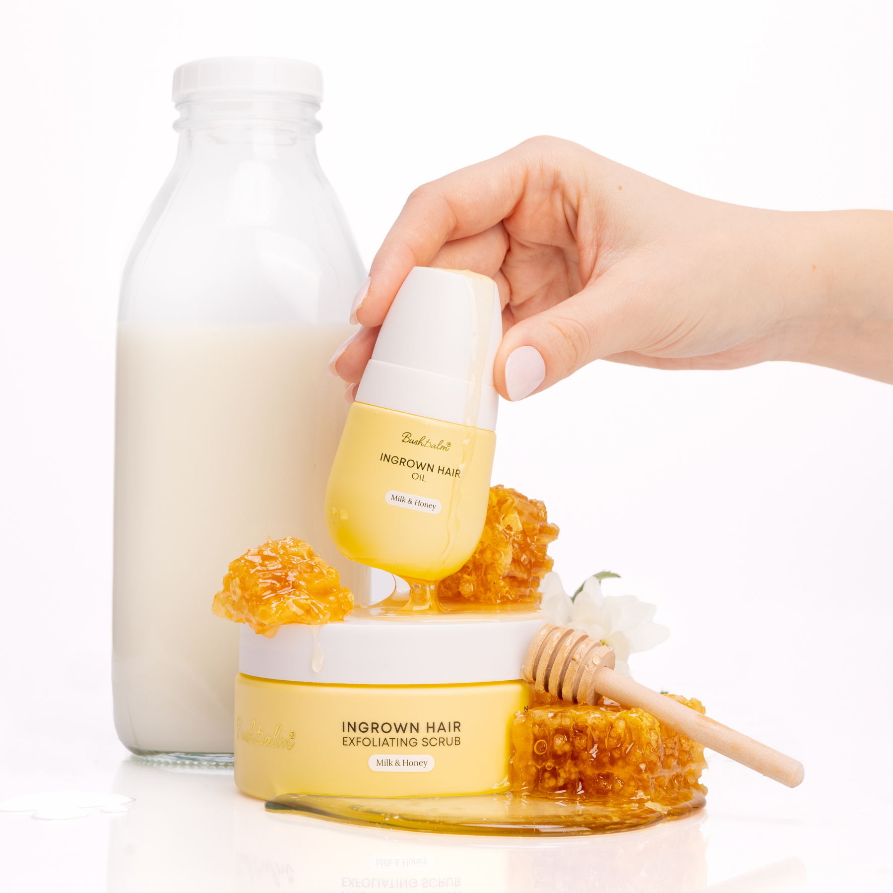 A hand holds milk and honey ingrown hair oil over a scrub, with a milk bottle and honeycomb.