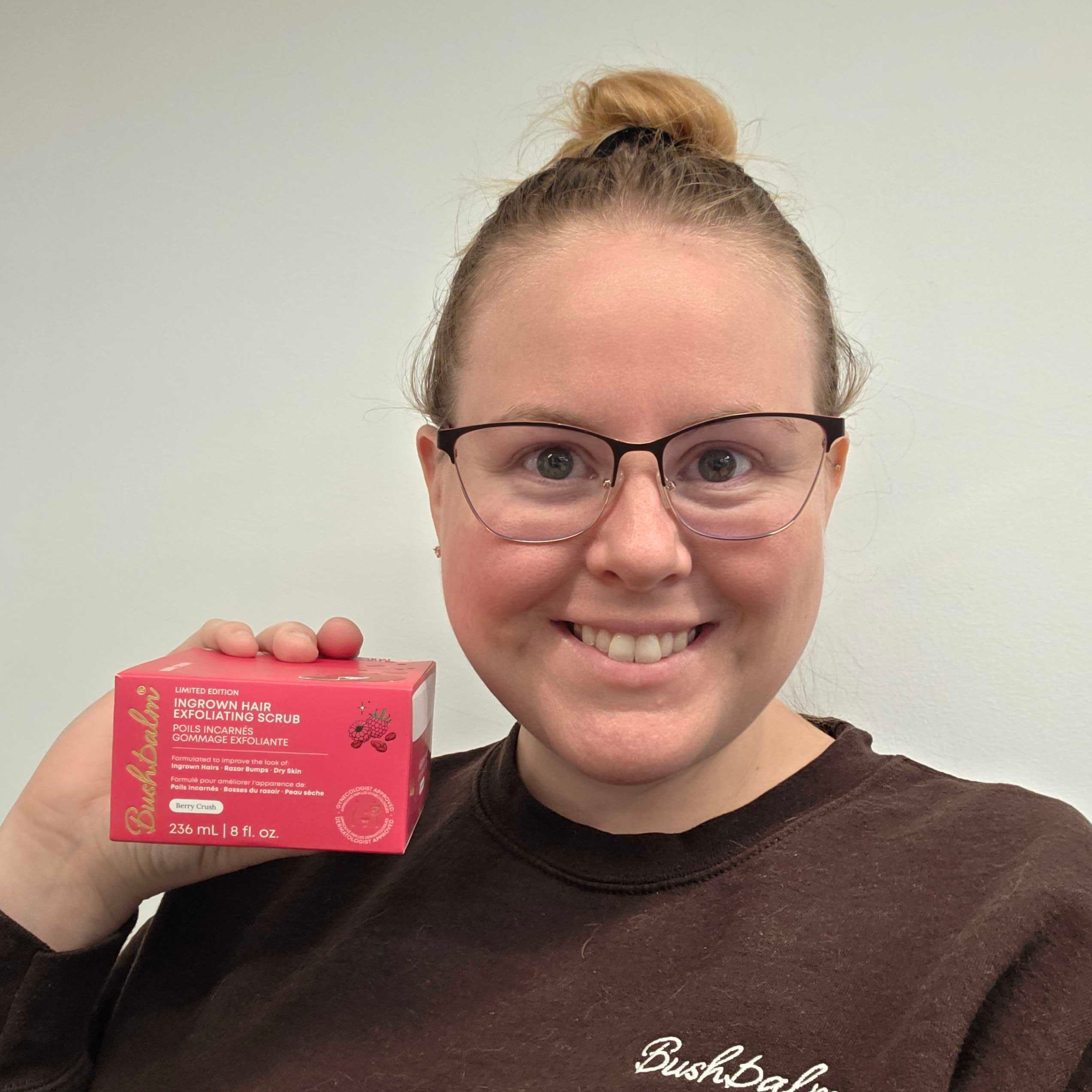 Person holding a box of ingrown hair exfoliating scrub, smiling against a plain background.