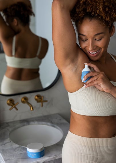 A smiling woman in a white top applies a product to her underarm in a bathroom.