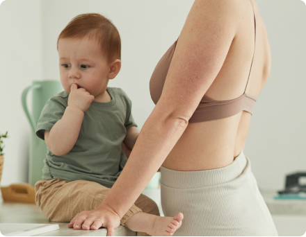 A baby sitting on a counter next to an adult in a kitchen.