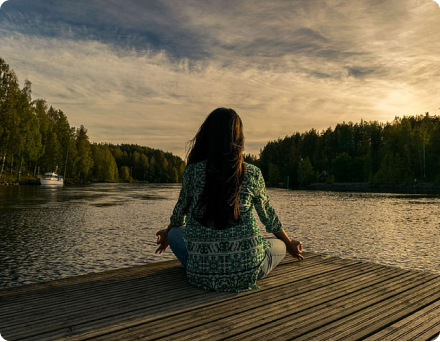 Person meditating on a wooden dock by a lake during sunset.