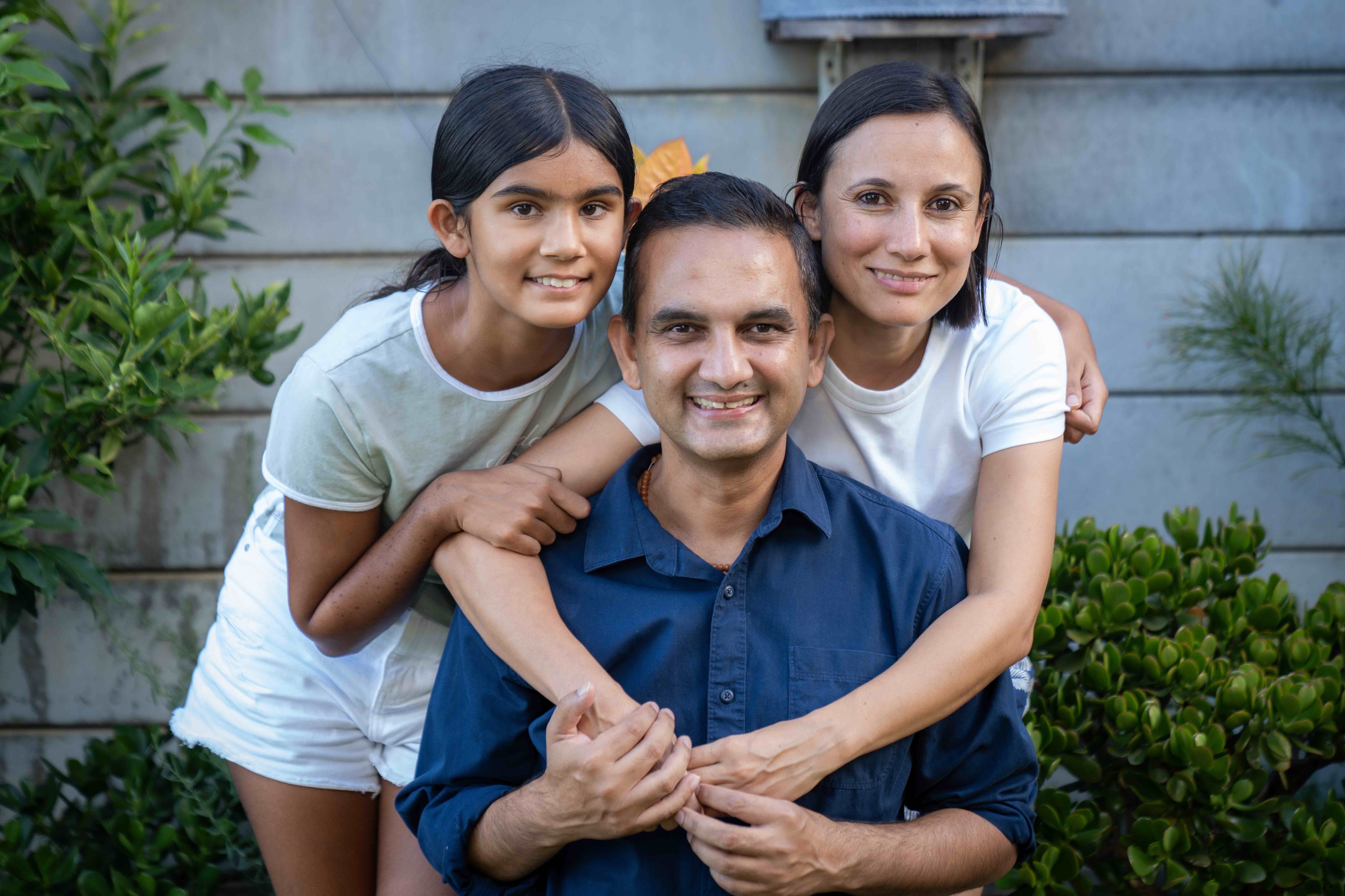 A smiling man, woman, and young girl pose for a photo outdoors while hugging.