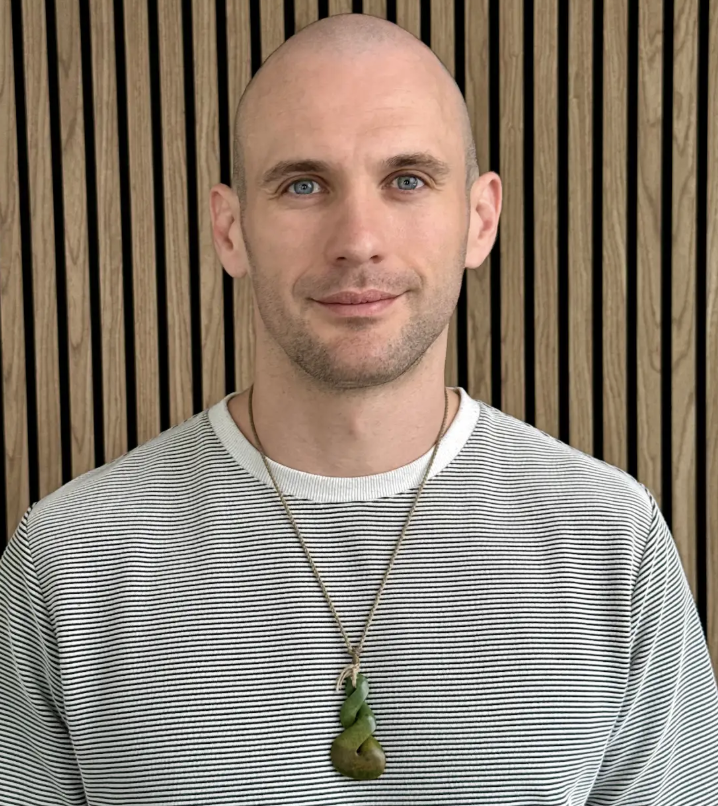 A bald man with blue eyes wearing a striped shirt and a green pendant necklace against a wood-slatted wall.