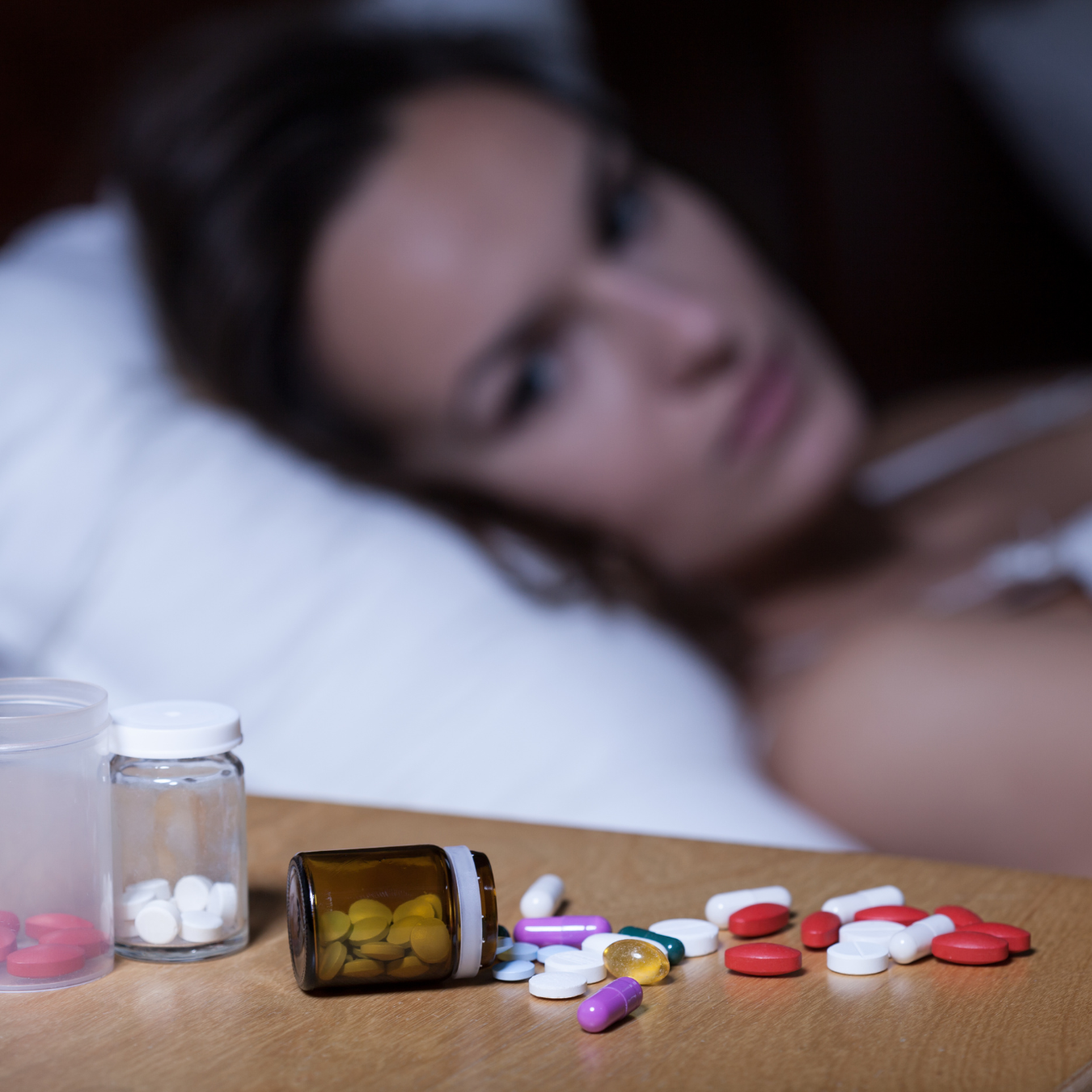 Pills scattered on a table with a person lying in the background.