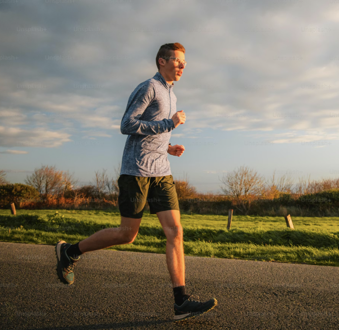 A young man with glasses jogs on a paved road next to a grassy field at sunset.