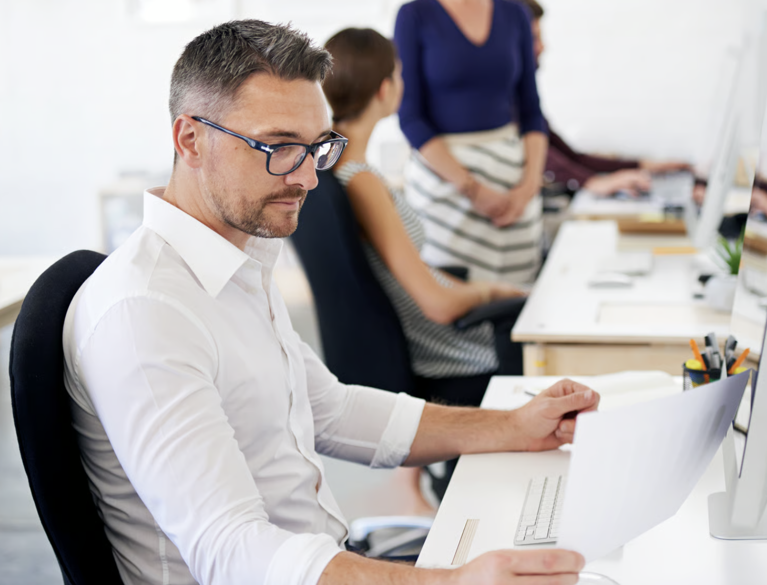 A man with glasses in a white shirt sits at a desk in an office, holding a paper.