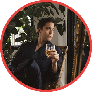 Person in a dark robe holding a glass, sitting by a window with plants.