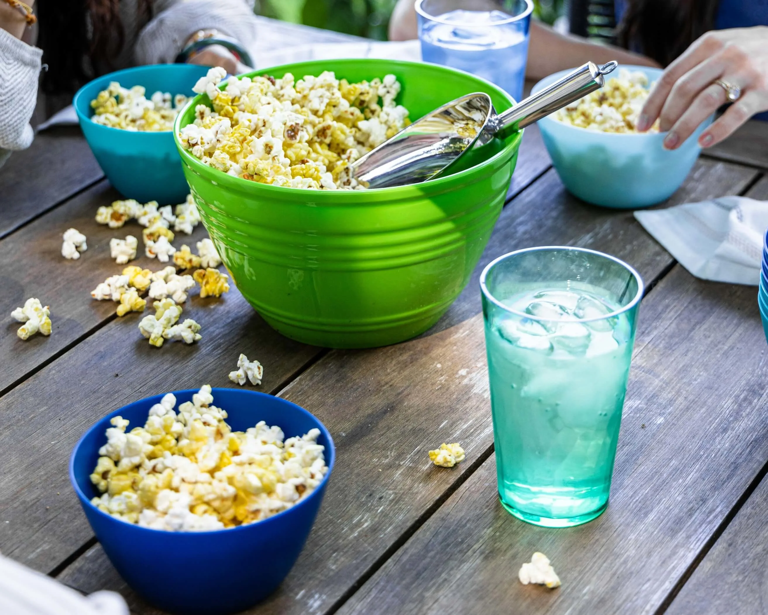 People share popcorn from colorful bowls on a wooden table with drinks.